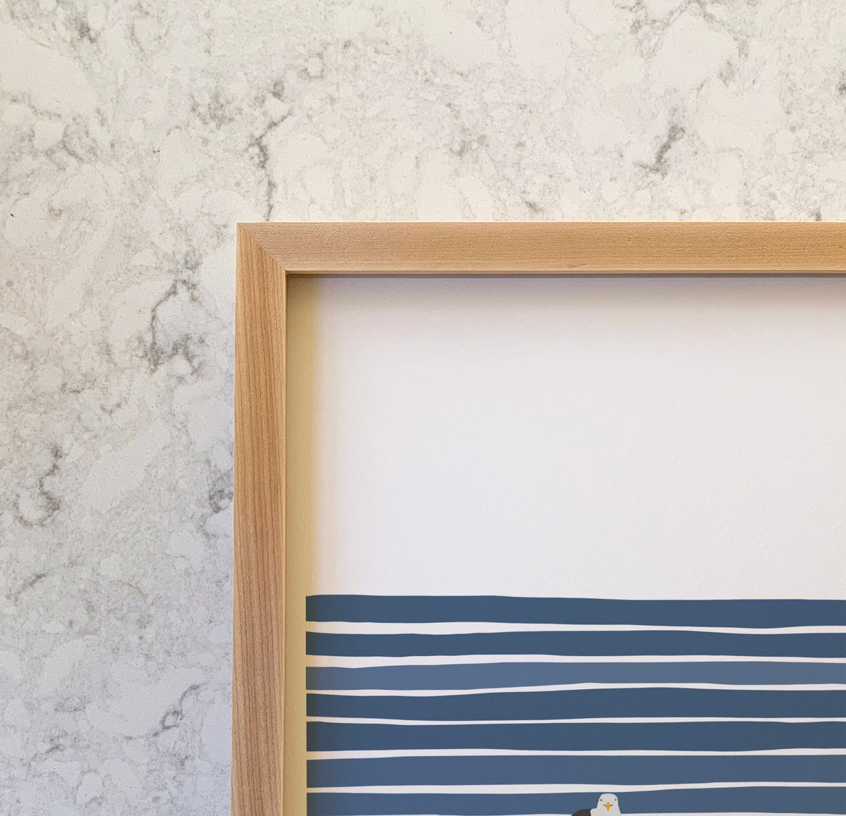 a closeup of the top left corner of a framed print by artist jorey hurley. the frame is solid wood, specifically maple, and is light in color. the visible portion of the art shows nautical navy blue and white stripes and part of a seagull.