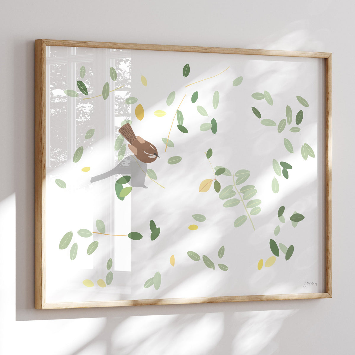 A framed art print featuring a minimalist illustration of a brown bird perched on a branch with green and yellow leaves, displayed on a white wall with soft window shadows.