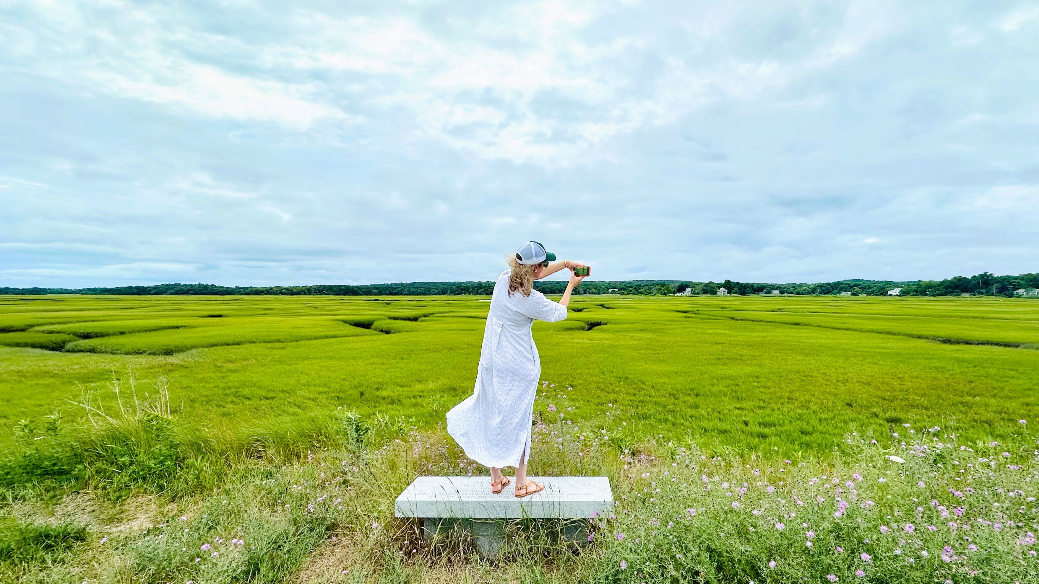 Jorey Hurley in a white dress standing on a bench in a green field with a cloudy sky - taking photographs as inspiration for her nature inspired artwork.