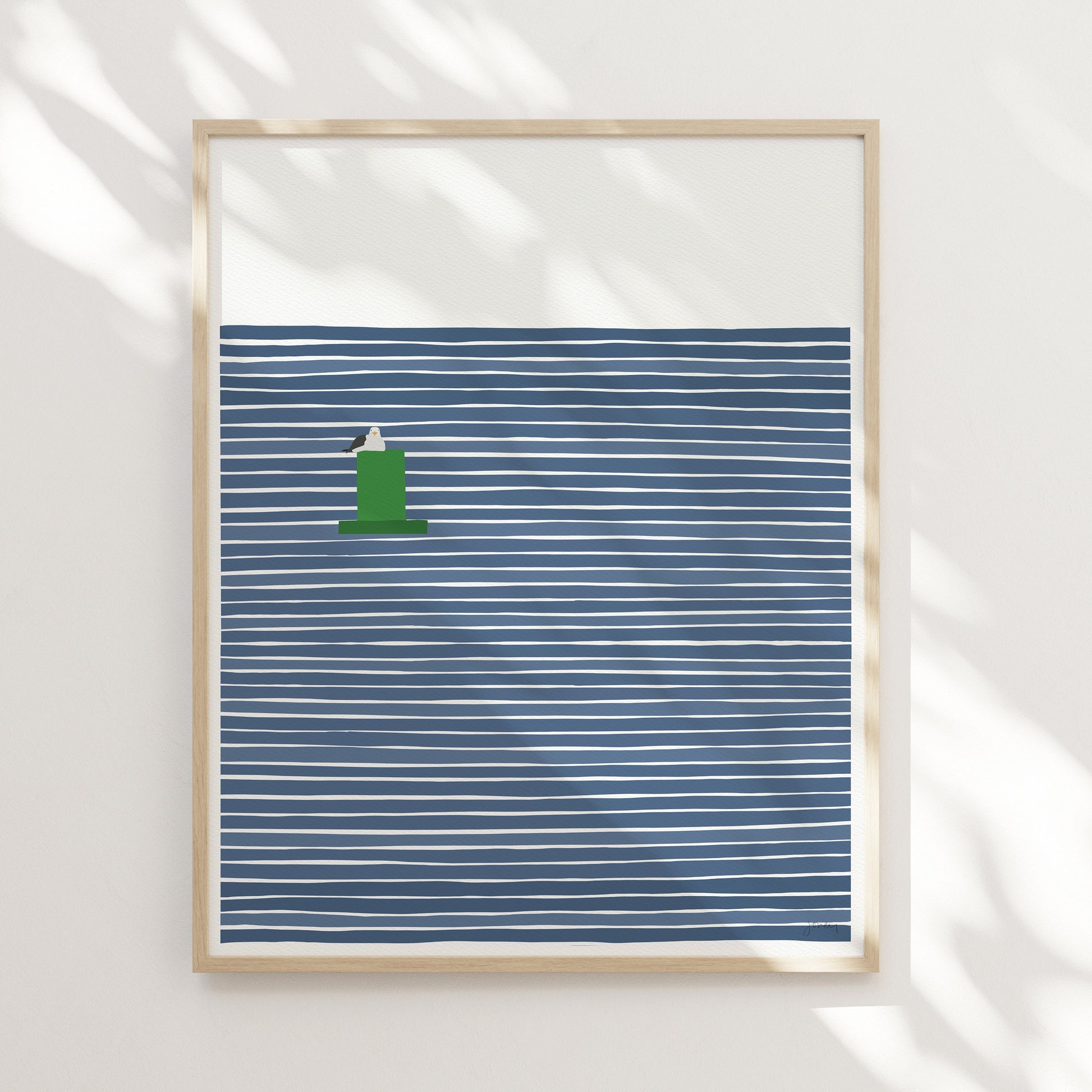a framed print by artist Jorey Hurley shown hung on a wall with light falling across it. the print is of a seagull perched on a buoy on a background of navy blue and white stripes. The frame is light wood colored.